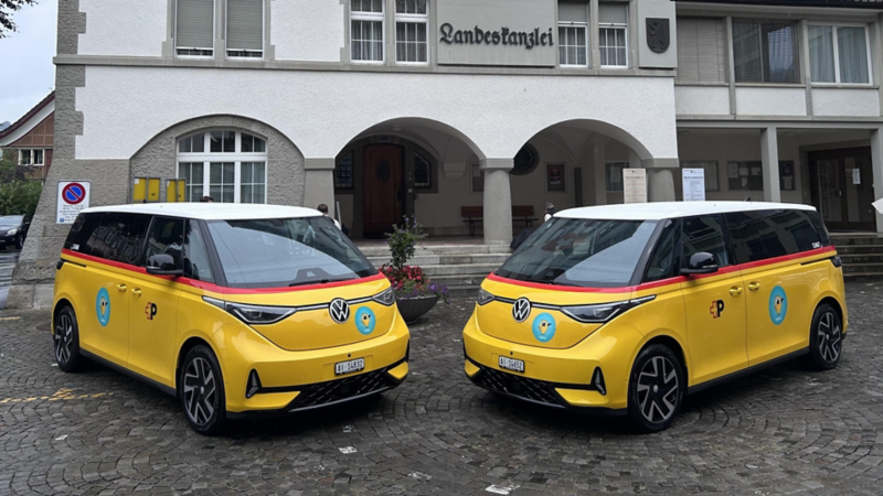 Zwei gelbe VW ID. Buzz im PostAuto-Design stehen nebeneinander auf einem Platz vor der «Landeskanzlei». Die Fahrzeuge tragen rote Zierstreifen, weisses Dach und das PostAuto-Logo