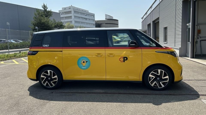 Seitliche Ansicht eines gelben VW ID. Buzz im PostAuto-Design auf einem Parkplatz. Auf der Schiebetür ist ein rundes Symbol mit Vogelmotiv zu sehen, daneben das PostAuto-Logo.