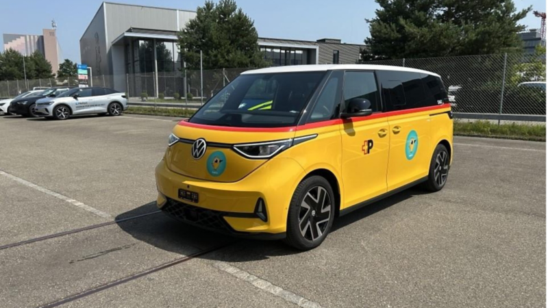 Front-seitliche Aufnahme eines gelben VW ID. Buzz im PostAuto-Design auf einem Parkplatz. Das Fahrzeug zeigt den typischen roten Streifen, das PostAuto-Logo und das runde Vogelmotiv.