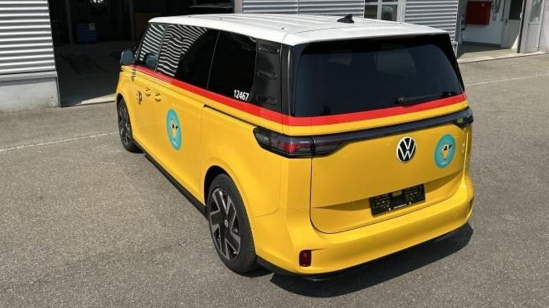Rück-seitliche Ansicht eines gelben VW ID. Buzz im PostAuto-Design mit weissem Dach. Das Fahrzeug steht vor einem Gebäude, sichtbar sind das runde Vogelmotiv und das durchgehende Rücklichtband