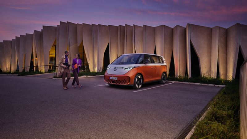 Volkswagen ID. Buzz People orange et blanc stationné sur un parking avec un couple qui marche à côté