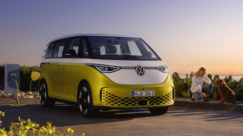 Volkswagen ID. Buzz jaune et blanc stationné sur un parking en charge. Une femme et un chien jouent sur le côté du véhicule. 