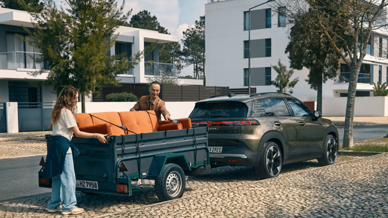 A man and woman are loading items into a trailer attached to the back of a green VW ID. Cross