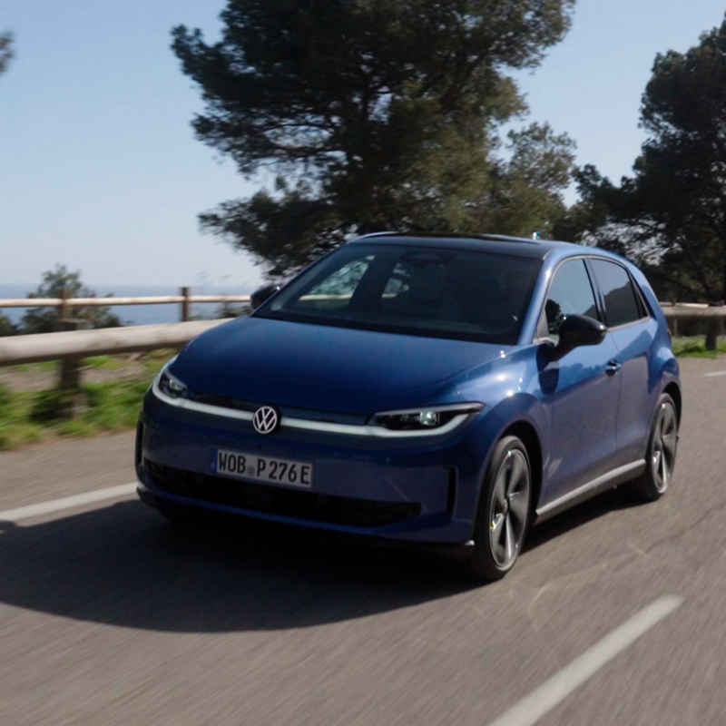 Front view of a blue Volkswagen ID.3 hatchback driving along a sunny road next to trees