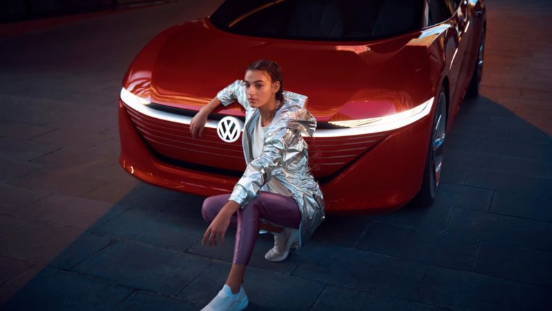 Chica con un chaleco plateado con el brazo encima de un Volkswagen ID. Vizzion rojo.