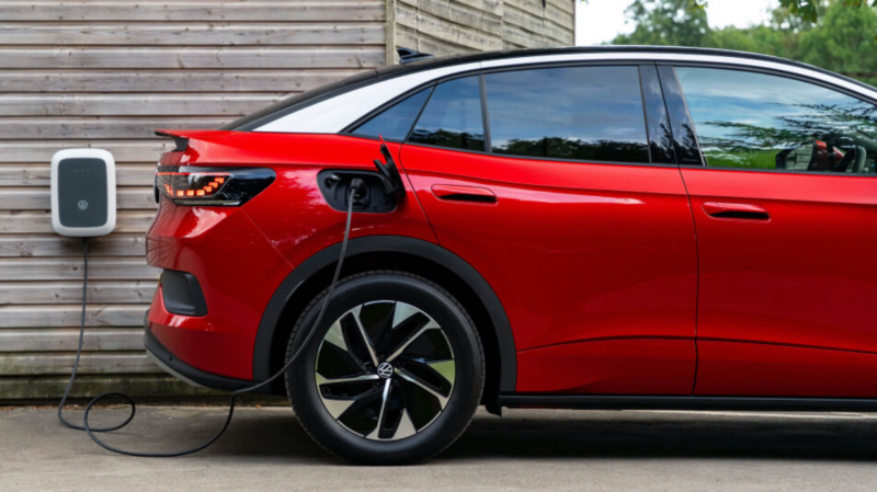 A red electric SUV is parked and plugged into a Volkswagen wallbox charger mounted on a wooden wall.