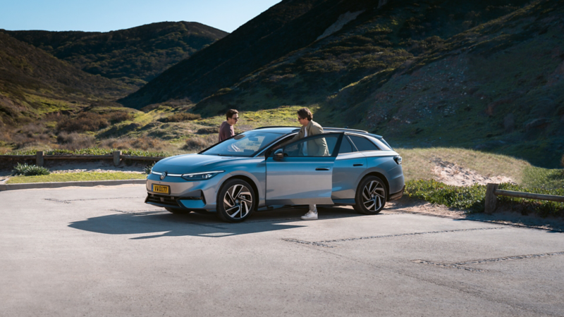 Deux hommes se tiennent à côté d'une Volkswagen ID.7 Tourer bleu clair métallisé. Le premier homme est assis au volant tandis que le second, debout, ouvre la portière avant passager. La voiture est garée sur un parking avec des collines et une végétation côtière en arrière-plan.