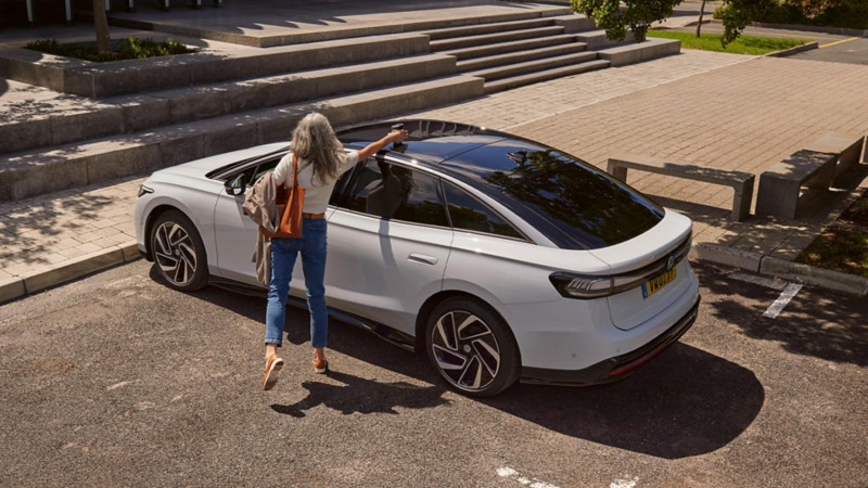 Une ID.7 blanche est garée, et la femme reprend le café déposé sur le toit de la voiture.