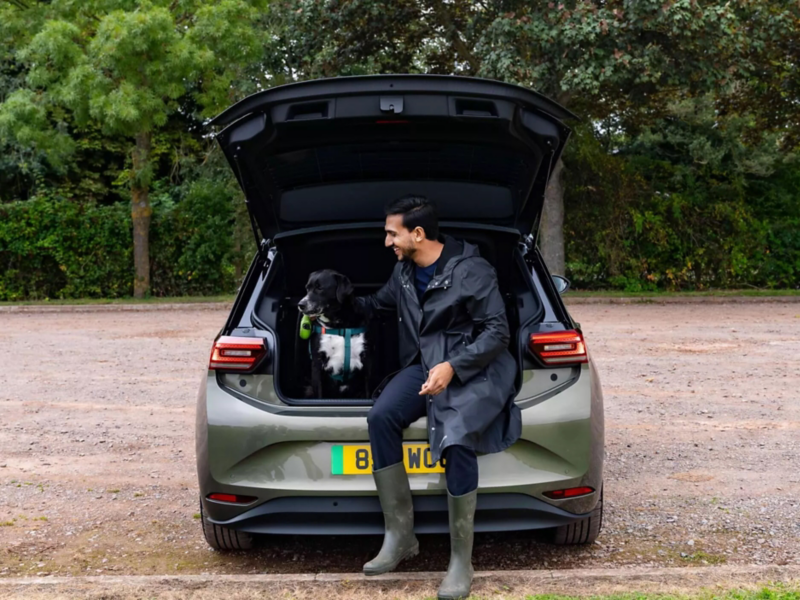 A man in a raincoat and boots sits in the back of a VW ID.3 with an open trunk, petting a black dog.