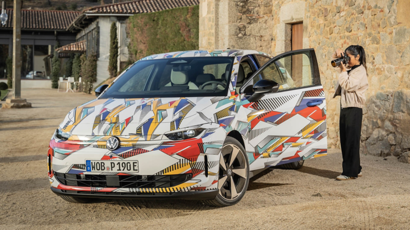 Eine Frau fotografiert einen bunt folierten Volkswagen ID. Polo, dessen Fahrertür geöffnet ist, vor einem historischen Gebäude.