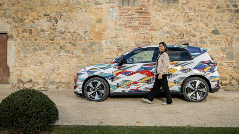 Eine Frau geht an einem bunt folierten Volkswagen ID. Polo vorbei, der vor einer historischen Steinmauer parkiert ist.