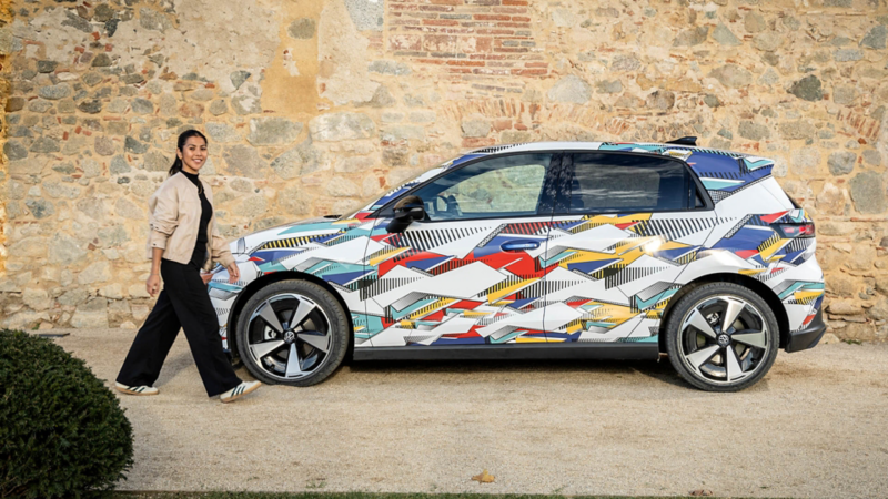 Seitenansicht eines bunt folierten Volkswagen ID. Polo, daneben geht eine Frau entlang einer Steinmauer.