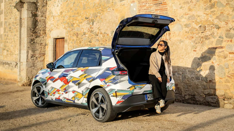 Eine Frau sitzt auf der offenen Heckklappe eines bunt folierten Volkswagen ID. Polo vor einer historischen Steinwand.