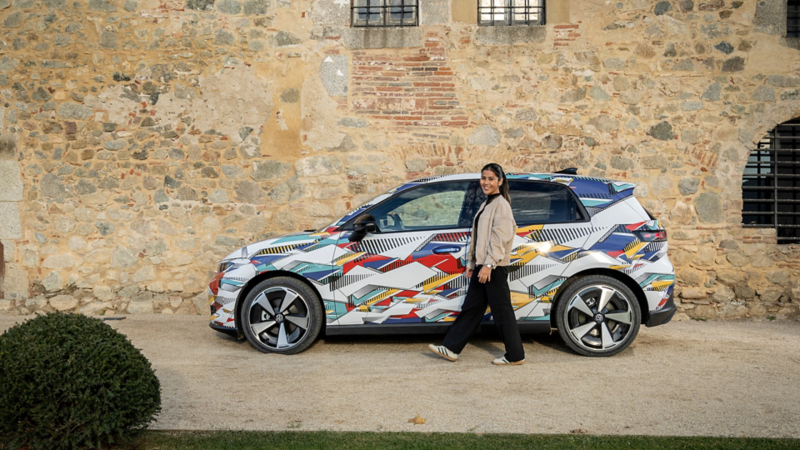 Eine Frau geht an einem bunt folierten Volkswagen ID. Polo vorbei, der vor einer historischen Steinmauer parkiert ist.