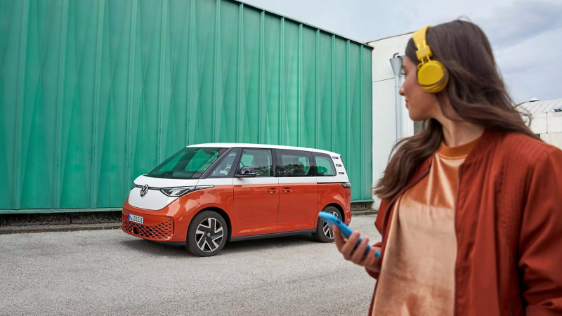 An orange and white Volkswagen ID. Buzz electric van parked in front of a turquoise wall, with a woman wearing yellow headphones in the foreground.