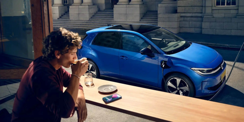 A man is sitting in a cafe, through an open window the blue VW ID. Polo can be seen charging at a charging station.