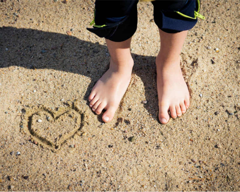 Person's feet on beach with a heart drawn in sand.