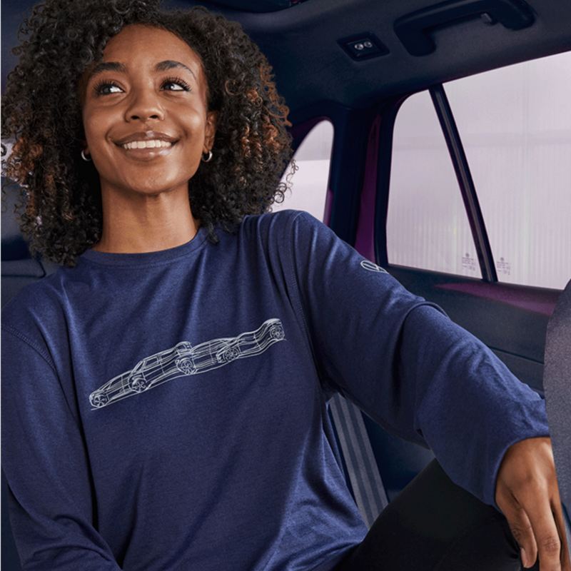 The image depicts a smiling woman with curly hair, sitting in a vehicle. She is wearing a long-sleeved dark blue shirt with a design of a car outline on the front. The interior of the vehicle is visible, with seats and windows in the background. The woman appears comfortable and relaxed, suggesting a positive experience within the car.