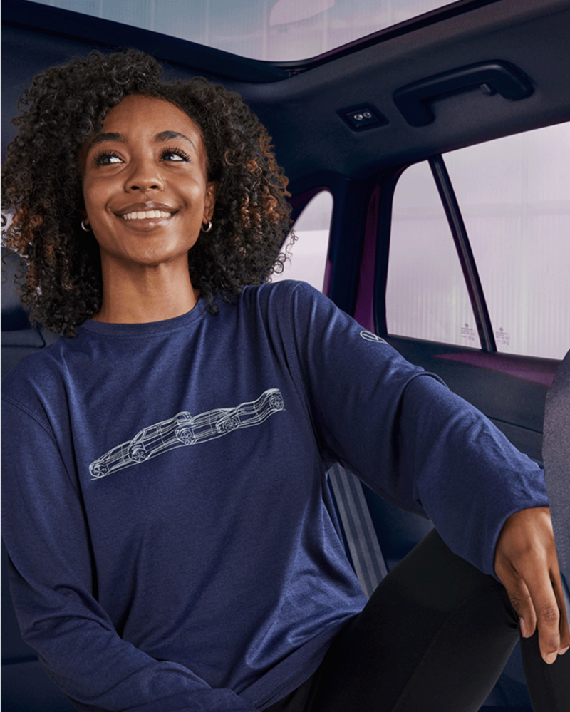 The image depicts a smiling woman with curly hair, sitting in a vehicle. She is wearing a long-sleeved dark blue shirt with a design of a car outline on the front. The interior of the vehicle is visible, with seats and windows in the background. The woman appears comfortable and relaxed, suggesting a positive experience within the car.