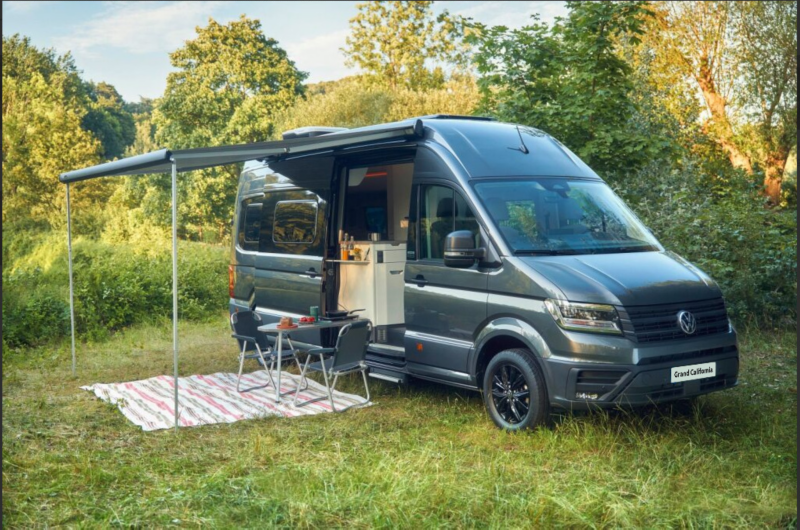 Volkswagen Grand California camper van parked at a campsite with outdoor chairs and extended awning.
