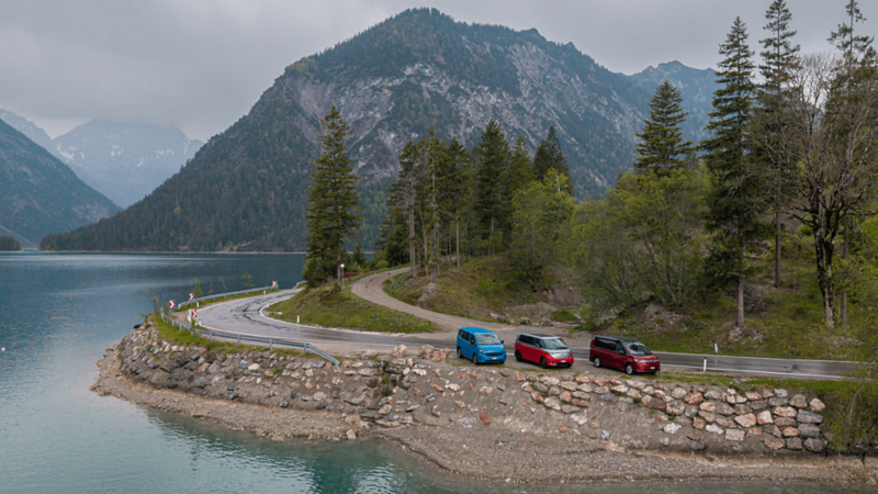 Tre modelli VW Bus sono parcheggiati su una strada curva lungo il lago con vista sulle montagne.