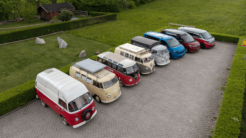 Sette VW Bus di diverse generazioni sono parcheggiati in fila su un posteggio.