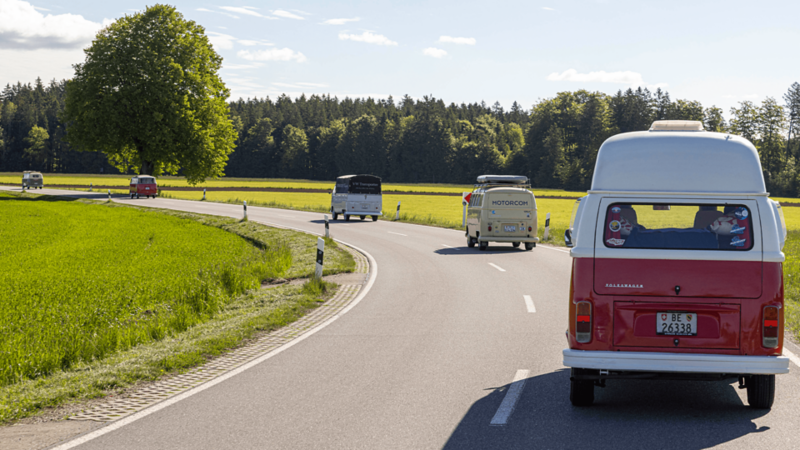 Plusieurs VW Bus classiques roulent en file sur une route de campagne.