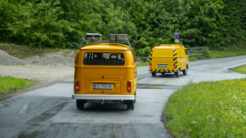 Deux VW Bus jaunes roulent dans un virage sur une route de campagne mouillée.
