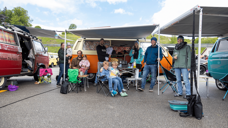Gruppo con bambini e VW Bus in campeggio sotto le tende da sole.