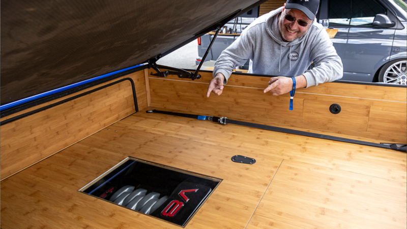 Un homme montre le moteur V8 intégré dans l’aménagement en bois d’un VW Bus.