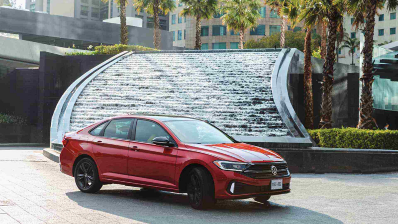 Jetta 2023, auto sedán de Volkswagen en color rojo, cerca de una fuente en un conjunto corporativo. 