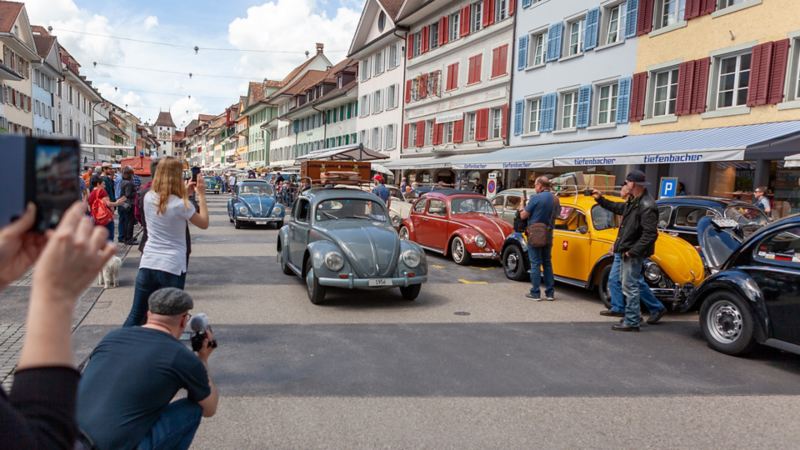 Mehrere VW Käfer fahren durch ein Dorf