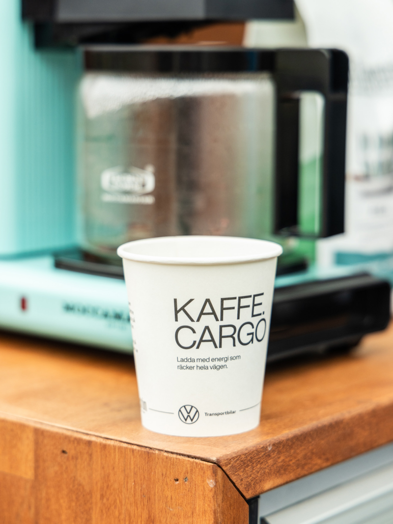 En pappmugg med trycket Kaffe Cargo står framför en turkos Moccamaster kaffemaskin