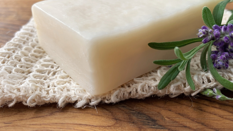 Nahaufnahme einer Kernseife, die auf einem Stück Stoff liegt, daneben liegt ein Stängel Lavendel.