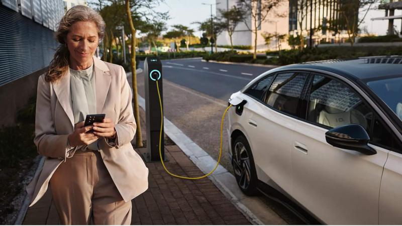 a woman looking at her phone next to a parked EV car that's charging