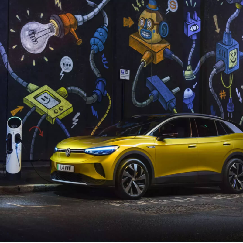 A vibrant yellow Volkswagen ID.4 electric SUV is parked on a city street in front of a colorful, electricity-themed graffiti wall art mural, next to a public charging station.