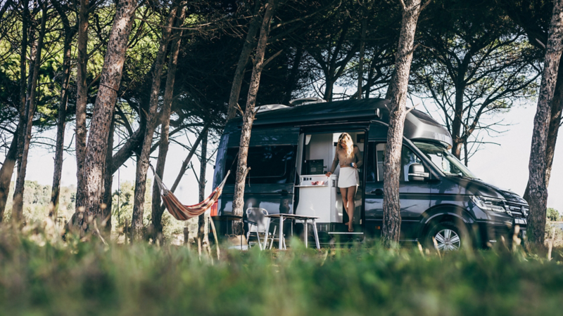 Volkswagen Camper auf einem Campingplatz im Wald, daneben eine Person am geöffneten Fahrzeug
