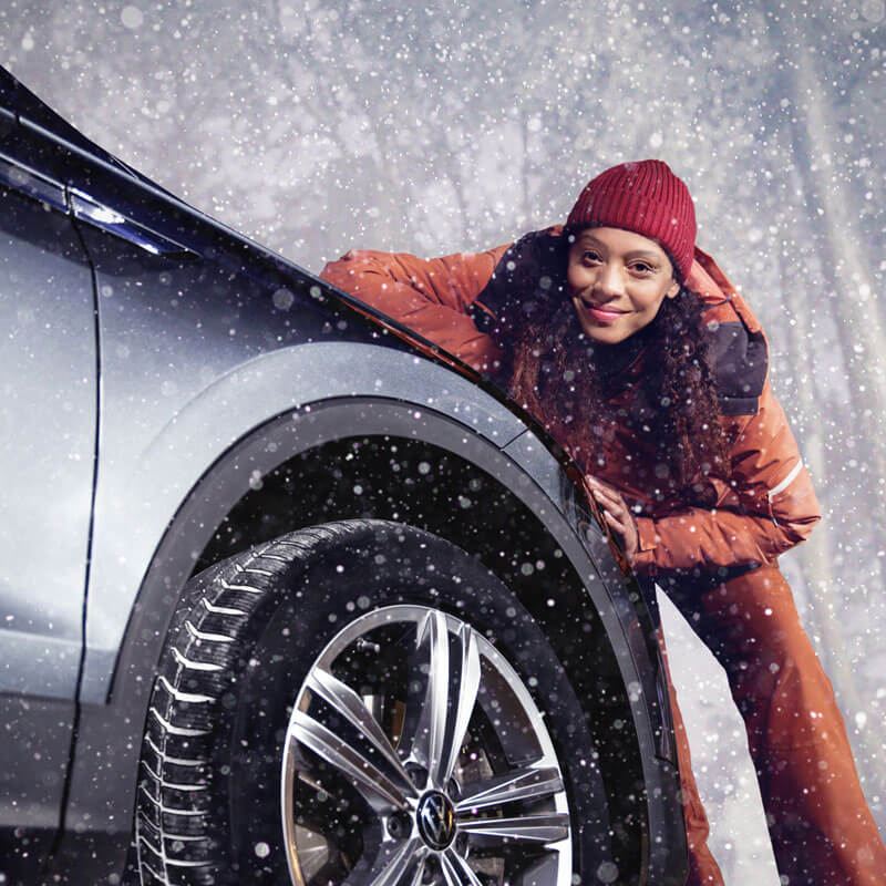 Une femme est penchée sur l'aile avant d'une Volkswagen chaussée de pneus hiver, la neige tombe.