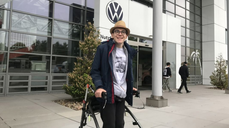 Luis Adam steht mit einer Gehhilfe vor einer VW Filiale.