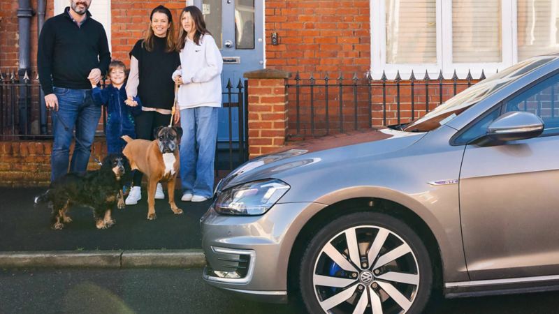 A family and their 2 dogs stand on the pavement outside their house next to a Golf GTE
