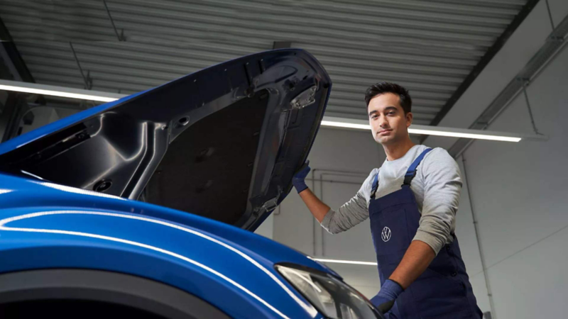 Técnico de Volkswagen abre el cofre de un SUVW de color azul, mientras mira a la cámara.