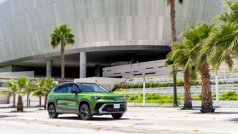 Taos de Volkswagen verde estacionado junto a palmeras, frente a un edificio moderno de fachada curva.
