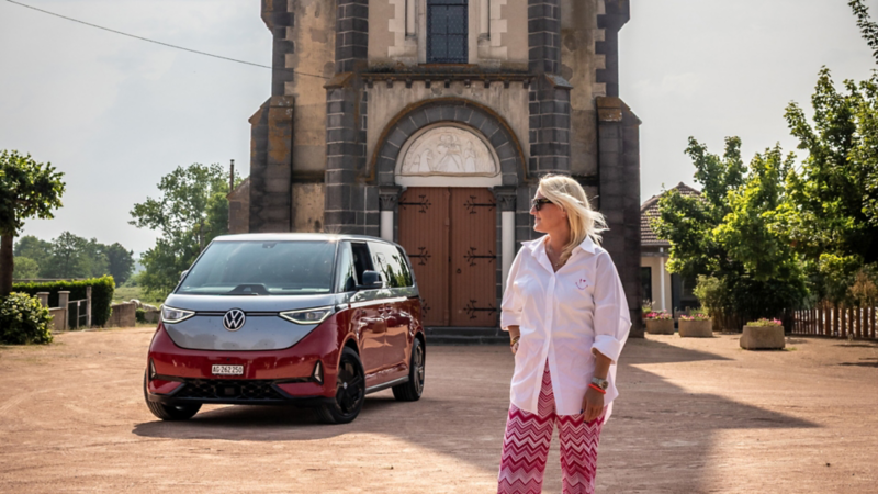 Une femme se tient devant une église historique, avec un VW ID. Buzz bicolore garé sur la place.