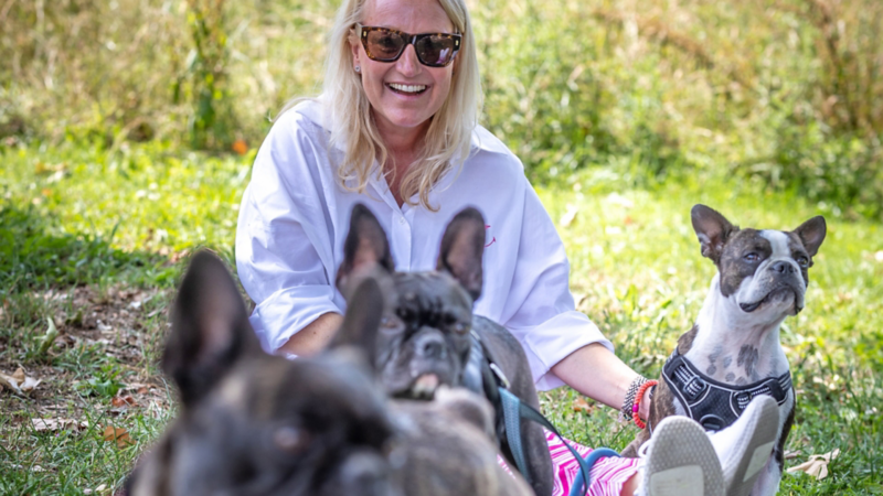 Une femme assise dans l’herbe rit, entourée de plusieurs petits chiens regardant vers la caméra.