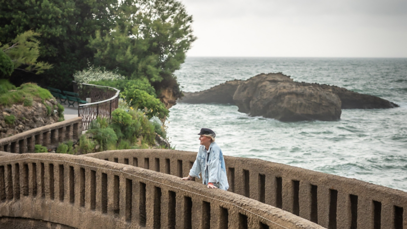 Eine Frau lehnt an einer Steinbrüstung und blickt aufs Meer, während Wellen gegen die Felsen schlagen.