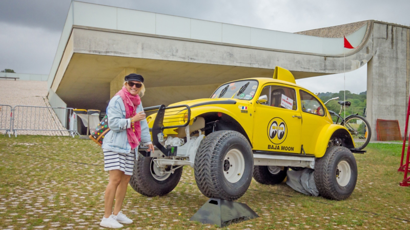 Eine Frau steht lächelnd neben einem gelben Baja-Käfer-Offroad-Fahrzeug auf einem Ausstellungsgelände.