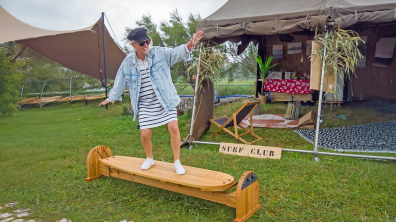 Une femme s’amuse à garder l’équilibre sur un simulateur de surf en bois devant une tente avec l’inscription « Surf Club ».