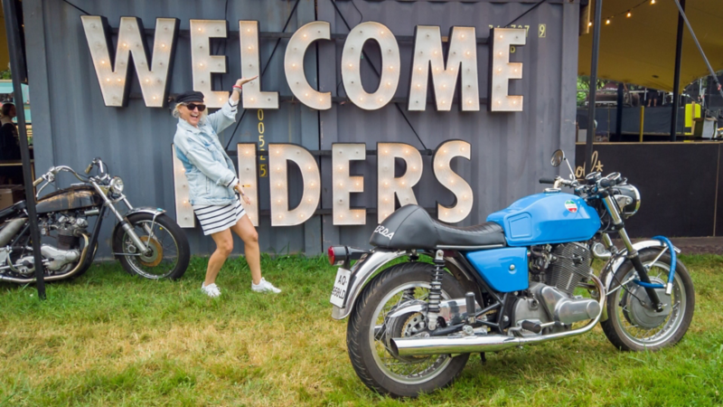 Une femme pose joyeusement devant une grande enseigne lumineuse « Welcome Riders », près de deux motos.