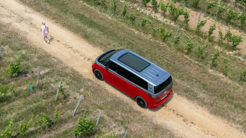 Ein VW ID. Buzz fährt auf einem Weg durch die Weinberge, während eine Frau vor dem Fahrzeug entlanggeht.