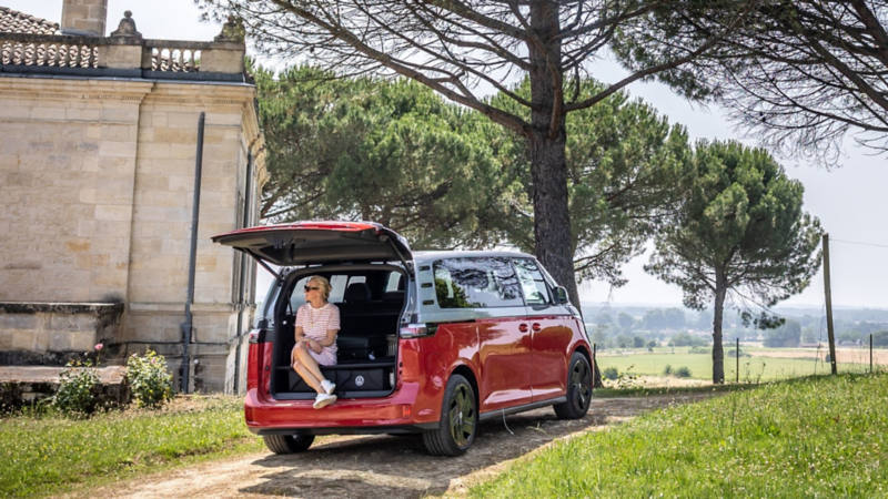 Eine Frau sitzt im geöffneten Kofferraum eines VW ID. Buzz, umgeben von Bäumen und ländlicher Landschaft.